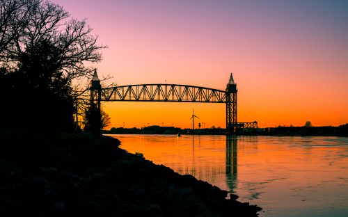 Silhouette of a bridge at sunset, reflecting on calm waters with trees and a wind turbine in the background.