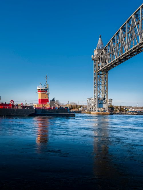 A tugboat navigates under a large bridge on a clear blue day, reflecting on the calm water below.