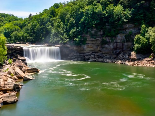 A serene waterfall cascades into a green river, surrounded by lush trees and rocky cliffs.