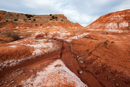 A rugged landscape of red and white clay hills under a cloudy sky, with a winding dry creek bed in the foreground.