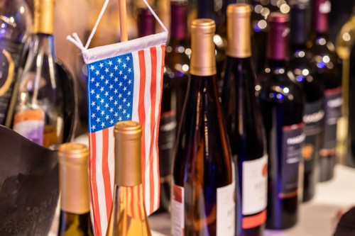 A small American flag hangs among various bottles of wine on a table.