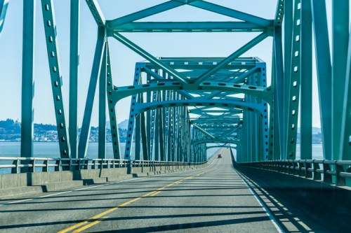 A view of a green steel bridge with a clear blue sky, leading over a road towards the horizon.