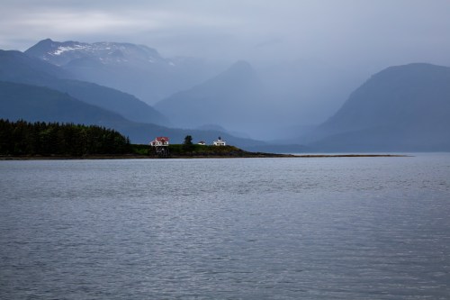 A serene landscape featuring mountains, a calm sea, and two distant houses on a shoreline under a cloudy sky.