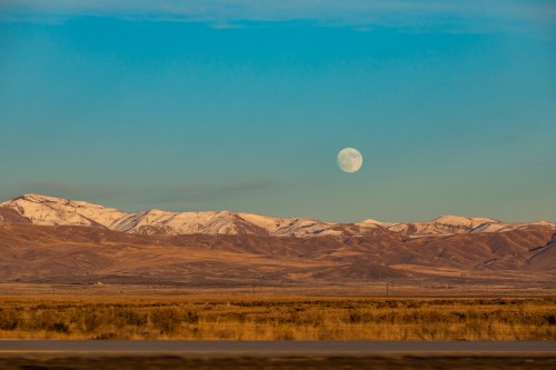 A serene landscape featuring snow-capped mountains under a clear sky with a large moon rising above the horizon.