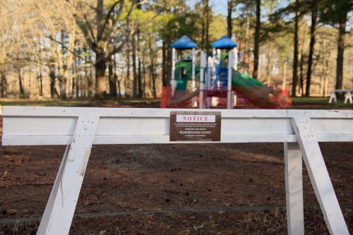 A closed playground sign on a barrier, with playground equipment visible in the background surrounded by trees.