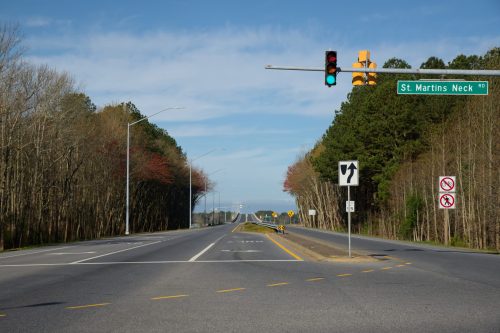 A wide road with traffic lights and a sign for St. Martins Neck Road, surrounded by trees under a clear blue sky.