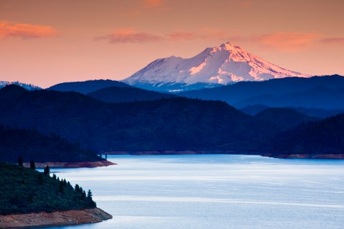 A serene lake reflects mountains at sunset, with a snow-capped peak in the background and lush hills surrounding the water.