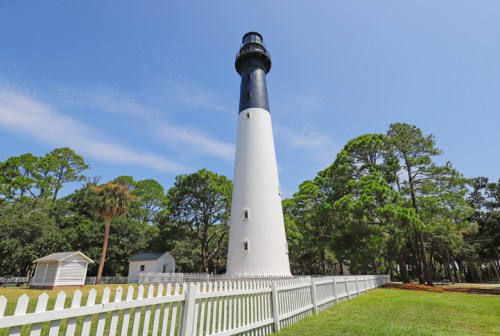 A tall lighthouse with a black top and white base, surrounded by green trees and a white picket fence.