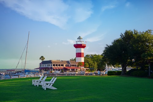 A red and white striped lighthouse beside a marina, with white chairs on green grass and a clear blue sky.