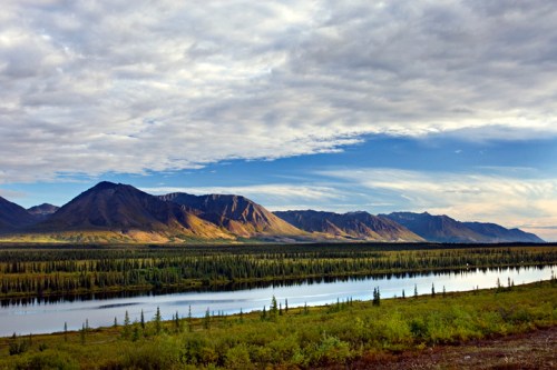 A serene landscape featuring mountains, a river, and lush greenery under a cloudy sky.