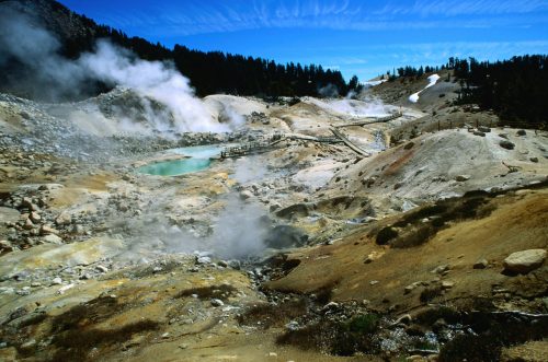 A geothermal landscape with steaming vents, a turquoise pool, and rocky terrain under a blue sky with wispy clouds.