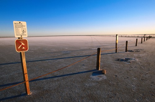 A vast, flat salt flat under a clear blue sky, marked by a fence and warning signs against trespassing.
