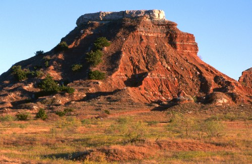 A rugged red rock formation rises against a clear blue sky, surrounded by sparse vegetation and golden grassland.
