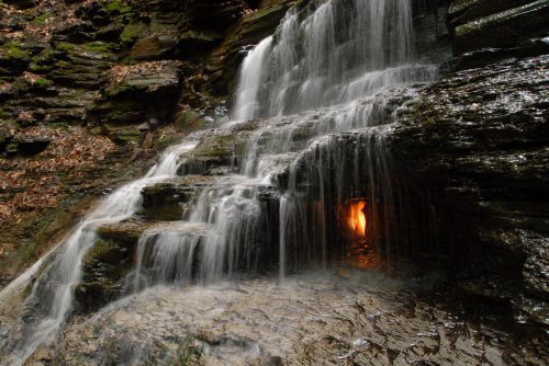 A serene waterfall cascades over rocky steps, with a warm glow emerging from a small opening in the stone.