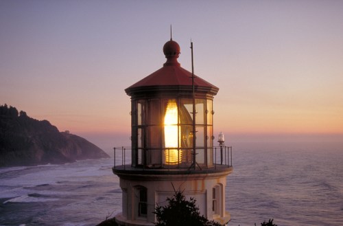 A lighthouse at sunset, with a glowing light and ocean waves in the background, surrounded by rugged cliffs.