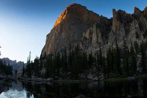 A serene mountain landscape at dawn, featuring a rocky peak, tall trees, and a calm reflective lake.