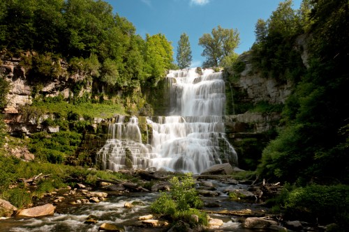 A cascading waterfall surrounded by lush greenery and rocky terrain under a clear blue sky.