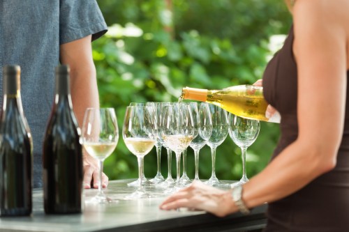 A person pours wine into glasses at an outdoor gathering, with bottles of wine in the background.
