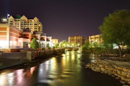 Nighttime view of a river with city buildings and lights reflecting on the water, surrounded by greenery.