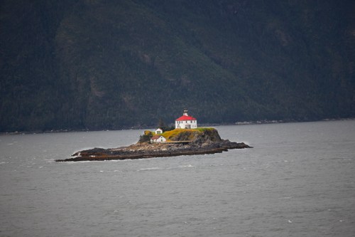 A small island with a lighthouse and a white building, surrounded by water and mountains in the background.