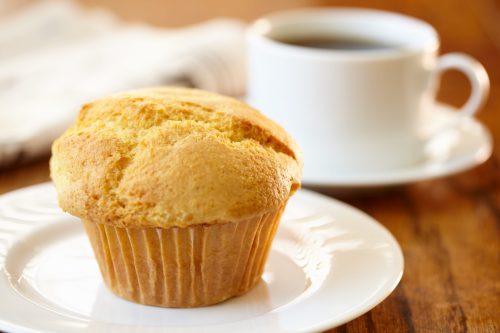 A golden muffin on a white plate next to a cup of coffee on a wooden table.