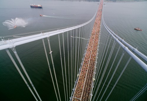Aerial view of a crowded bridge over water, with boats nearby and a ship spraying water in the background.