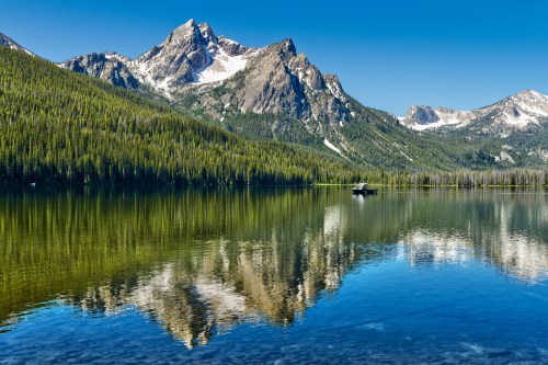 A serene lake reflects majestic mountains and lush forests under a clear blue sky.