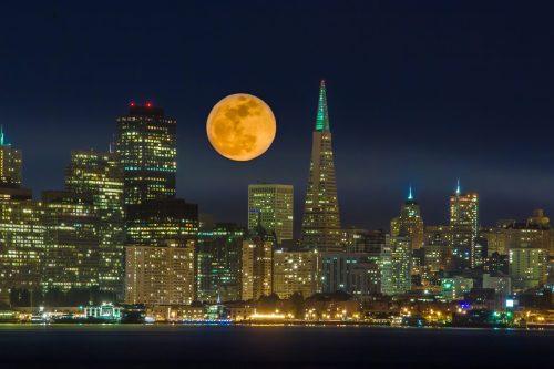 A vibrant city skyline at night with a large, glowing moon rising above tall buildings, including a spire.