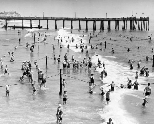 Black and white photo of a crowded beach with people swimming and playing in the water near a pier.