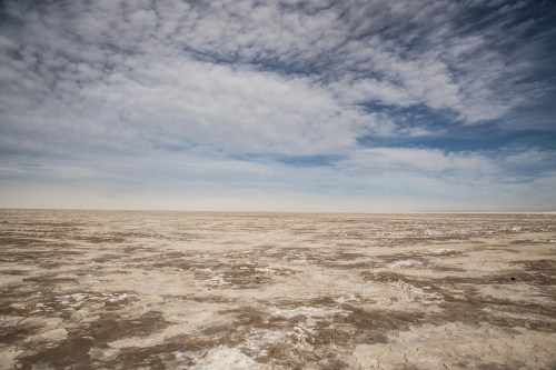 A vast, dry salt flat under a cloudy blue sky, with cracked earth stretching to the horizon.