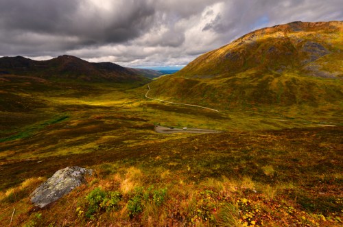A scenic view of rolling hills and valleys under a cloudy sky, with a winding road and patches of green and gold foliage.
