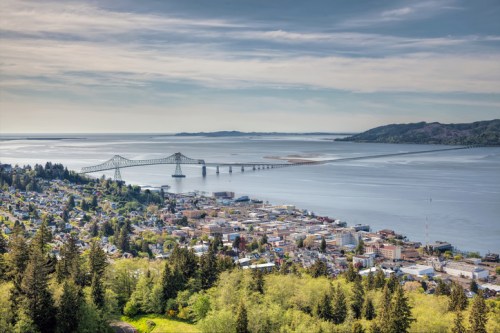 Aerial view of a coastal town with a bridge spanning a river, surrounded by lush greenery and distant hills.
