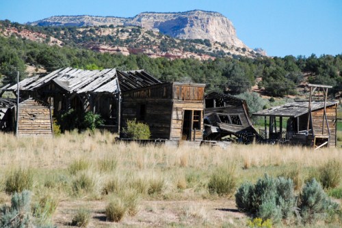 Abandoned wooden buildings in a grassy landscape, with a rocky cliff in the background under a clear blue sky.