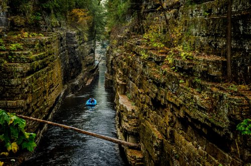A person in a kayak navigates a narrow, rocky canyon surrounded by lush greenery and autumn foliage.