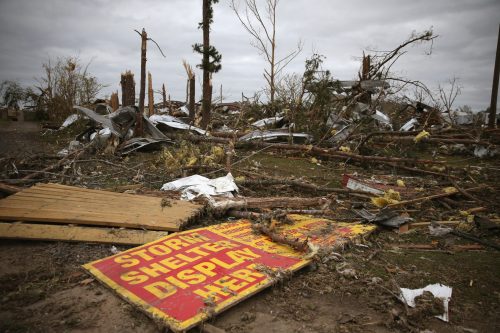 A damaged sign reading "STORM SHELTER DISPLAY HERE!" amidst debris and fallen trees after a storm.