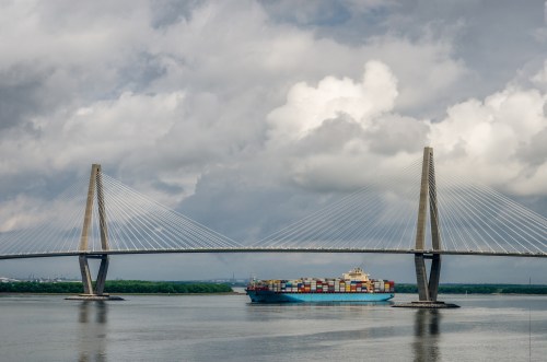 A large container ship passes under a modern cable-stayed bridge on a cloudy day.