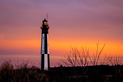 A lighthouse stands against a colorful sunset, surrounded by silhouettes of grass and shrubs.