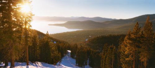 A panoramic view of a serene lake surrounded by mountains and evergreen trees, illuminated by a warm sunrise.