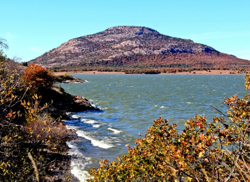 A serene lake with gentle waves, framed by trees, and a prominent mountain in the background under a clear blue sky.