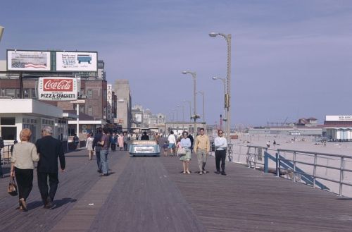 A busy boardwalk scene with people walking, vintage cars, and beachside buildings under a clear blue sky.