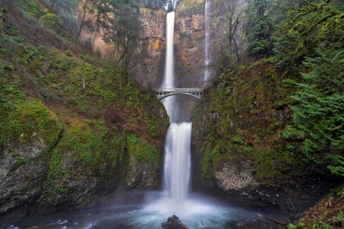 A stunning waterfall cascades down rocky cliffs, surrounded by lush greenery and a bridge spanning the water below.