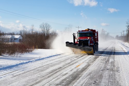 A red snowplow clears a snowy road under a bright blue sky, with trees and a house in the background.