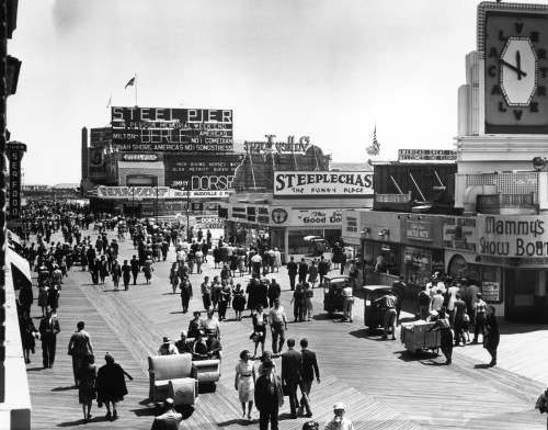 A busy boardwalk scene with crowds, shops, and signs, capturing a lively atmosphere in black and white.