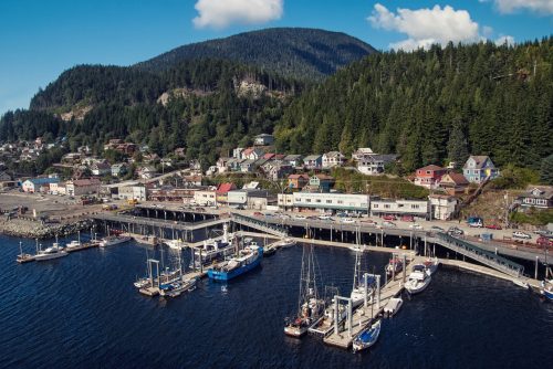 Aerial view of a coastal town with colorful houses, a busy dock, and boats moored in the water, surrounded by mountains.