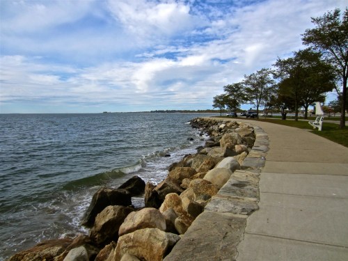 A scenic shoreline with a rocky edge, a paved path, and trees under a cloudy sky. Waves gently lap at the rocks.