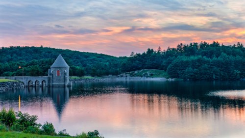 A serene lake at sunset, featuring a stone tower and lush green trees reflecting in the calm water.