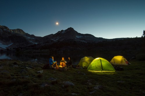 A serene campsite at night with three people around a fire, illuminated tents, and a mountain backdrop under a starry sky.