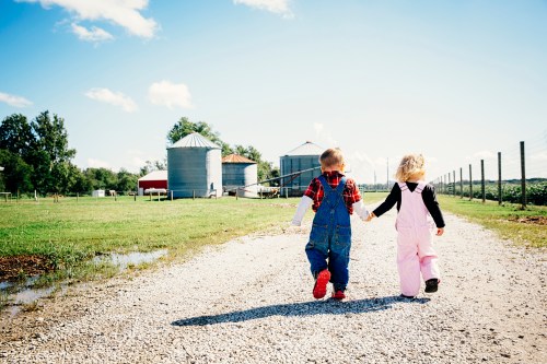 Two children in overalls walk hand in hand down a gravel path, with silos and a farm in the background.