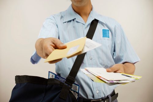A postal worker in a blue uniform holds out an envelope while carrying a bag filled with mail.