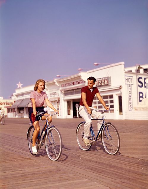 Two young people ride bicycles along a boardwalk, with shops and a pastel sky in the background.